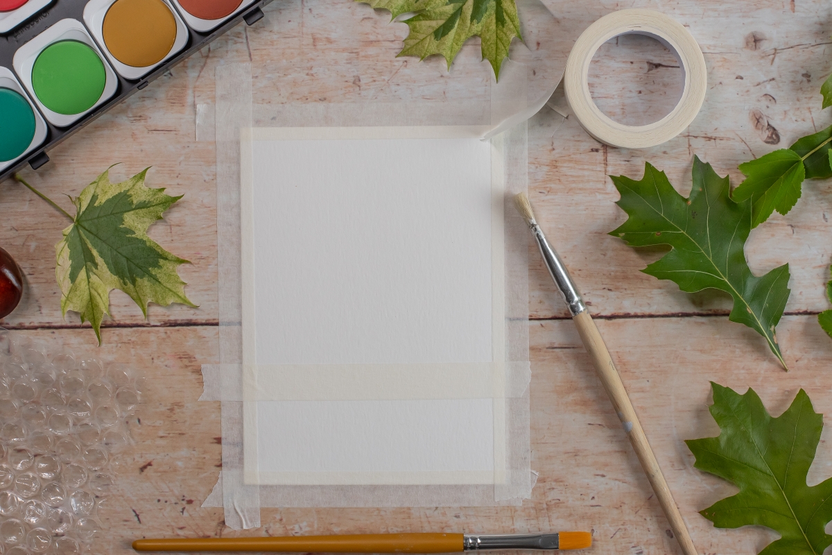 Ein Blatt Papier ist auf eine Unterlage geklebt - Pinsel, Tuschkasten und Bl&auml;tter liegen daneben.